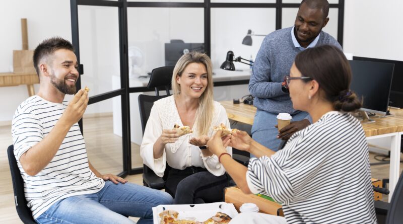 Quatro colaboradores sorridentes em ambiente de escritório moderno compartilhando uma pizza durante o intervalo, representando a satisfação com o benefício alimentação refeição empresa