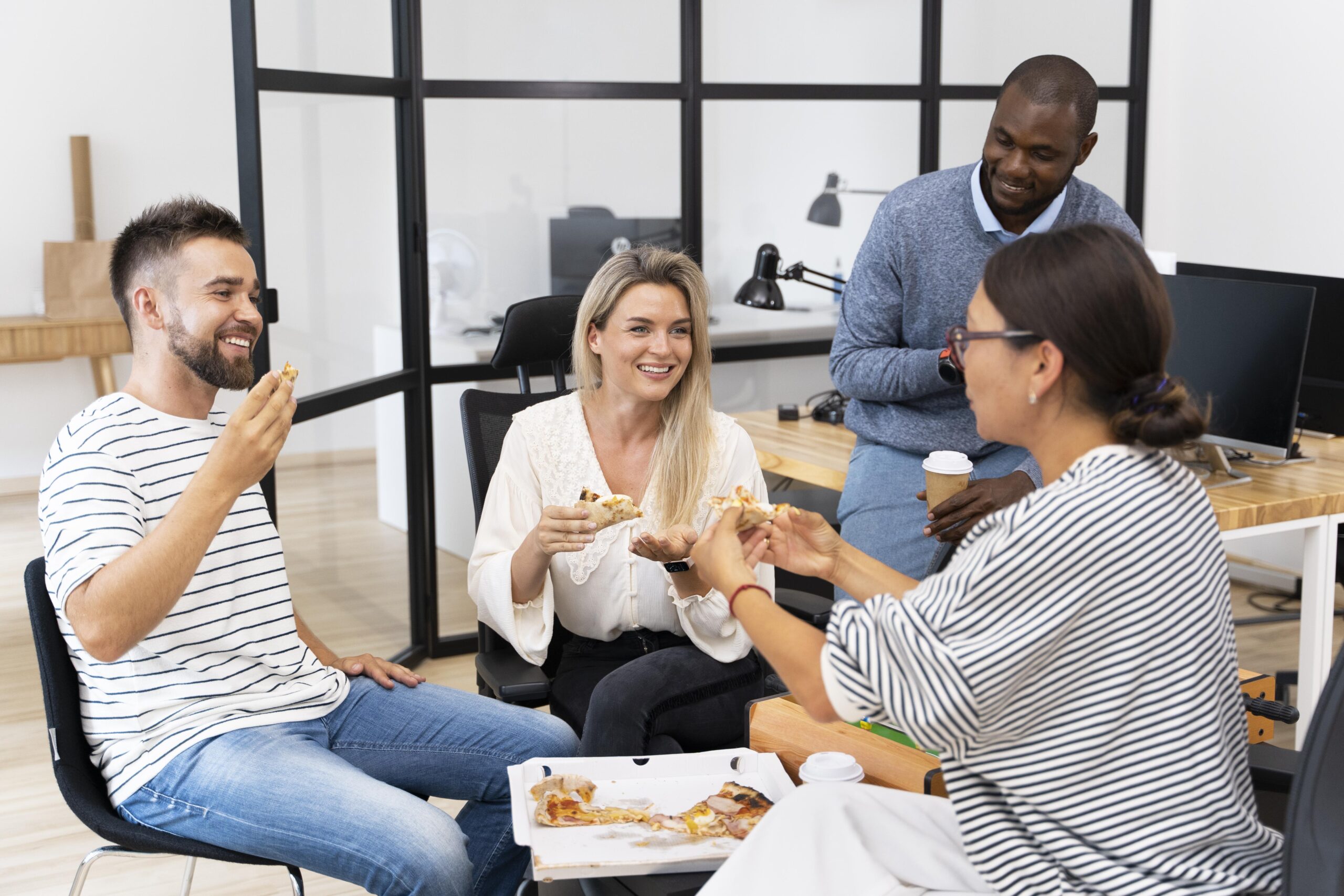 Quatro colaboradores sorridentes em ambiente de escritório moderno compartilhando uma pizza durante o intervalo, representando a satisfação com o benefício alimentação refeição empresa